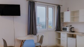 Een kleine kitchenette met een magnetron, waterkoker en kastjes, een aan de muur bevestigde televisie en een raam met gordijnen.