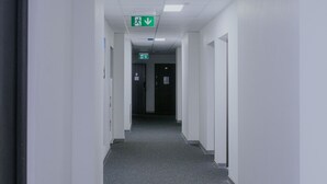 Een lange, smalle gang met witte wanden en een grijs vloerkleed. Aan beide kanten bevinden zich deuren en erboven hangt een groen bord met daarop een nooduitgang.'