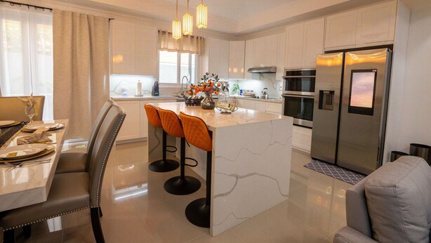 A modern kitchen with white cabinetry, a central island with bar stools, stainless steel appliances, and a dining area with a table set for four. - The Luxurious Chateau (Kingston)