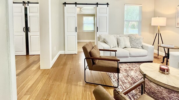 A modern living room with a white sofa, a brown armchair, and a round coffee table. There is a sliding door leading to another room and a window with blinds. - “1933” Sleeps 6,  King Beds, Spacious, 5 min walk to downtown Mt. Dora  (Mount Dora)