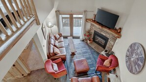 A spacious living room with a fireplace, a large TV, and a balcony with sliding doors. - Cozy 3-Level Condo w/ Loft Bedrooms w/ Fireplace (Brian Head)