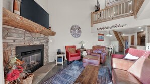 A spacious living room with a stone fireplace, leather sofas, a wooden coffee table, and a large clock on the wall. - Cozy 3-Level Condo w/ Loft Bedrooms w/ Fireplace (Brian Head)