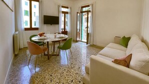 A living room with a sofa, a dining table with chairs, a television mounted on the wall, and large windows with curtains. - Splendid "True Venice Apartment" overlooking water (Venezia)