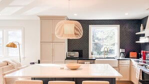 Et moderne køkken med en central ø, træbarstole, en hængende lampe og udsigt til det frie gennem et vindue.