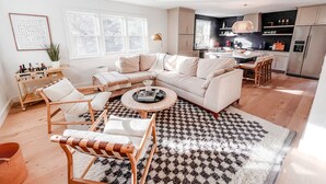 En moderne stue med sofa, stole og sofabord. Et køkken er synligt i baggrunden med skabe og hårde hvidevarer.