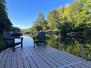 Une jetée en bois avec deux chaises Adirondack, une rivière paisible et des bateaux amarrés au loin.