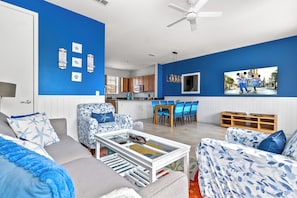 A living room with a blue wall, a ceiling fan, a dining area with a table and chairs, and a kitchen in the background. - Beachside Vibes |2BR/2BATH Townhouse | Near Disney (Kissimmee)