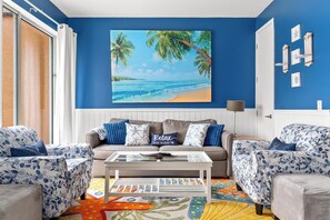 A living room with a blue wall, a beach-themed painting, a sofa with starfish pillows, and a colorful rug. - Beachside Vibes |2BR/2BATH Townhouse | Near Disney (Kissimmee)