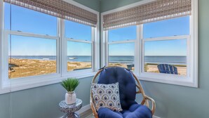 Una habitación con vista a la playa, una silla de mimbre con cojín y una planta pequeña sobre una mesita auxiliar.