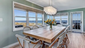 Un comedor con mesa de madera, sillas de mimbre, una lámpara de araña y ventanas amplias con vista a la playa.