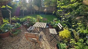 Un jardín con una mesa y sillas de madera, rodeado de plantas en macetas y un camino de grava.