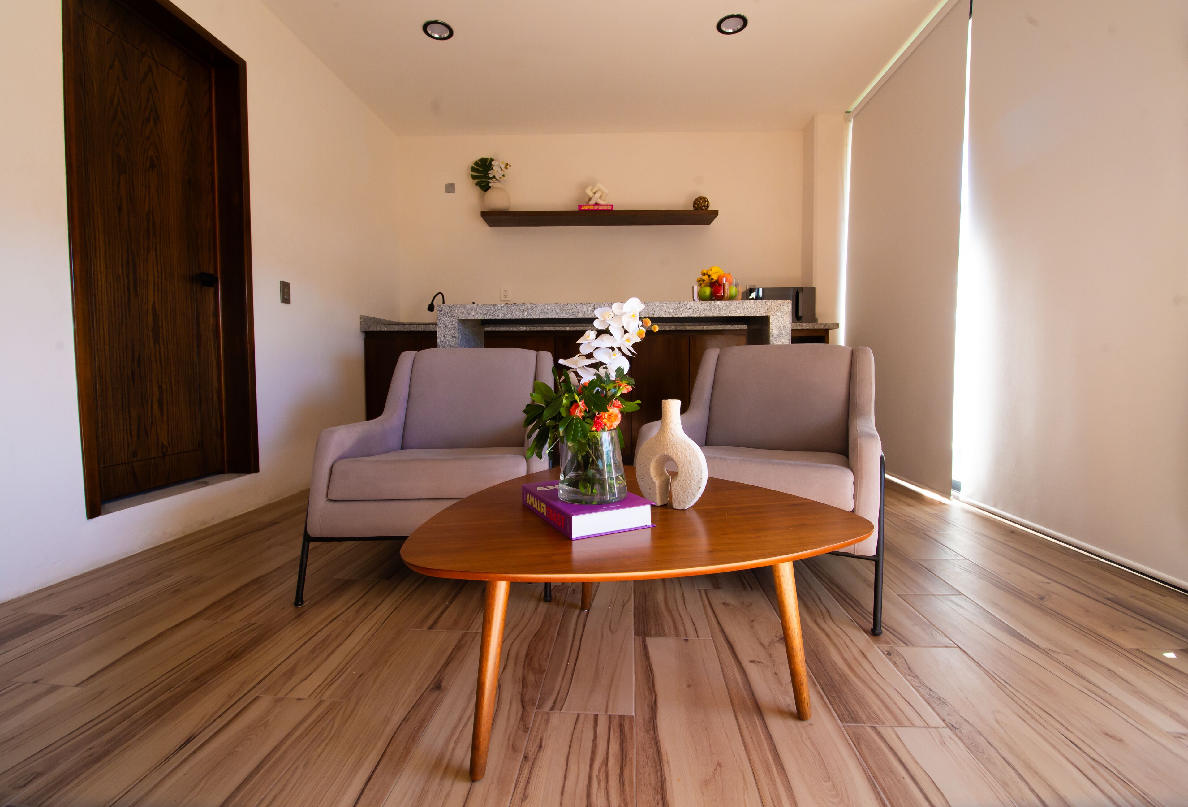 A modern living room with a wooden coffee table, two grey sofas, a kitchen in the background, and a wooden floor.
