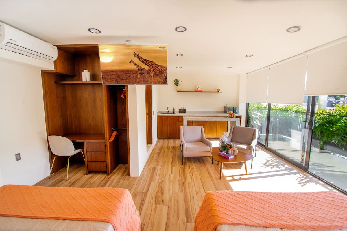 Una habitación de hotel moderna con un escritorio de madera, dos sillas, una cama con colcha naranja, un ventanal con vistas a zonas verdes y una zona de cocina.