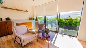 A modern living room with a sofa, a coffee table with decorative items, a kitchen area with a countertop and cabinets, and a large window with a view of greenery and buildings. - KUKO SUITES (Chilpancingo de los Bravo)