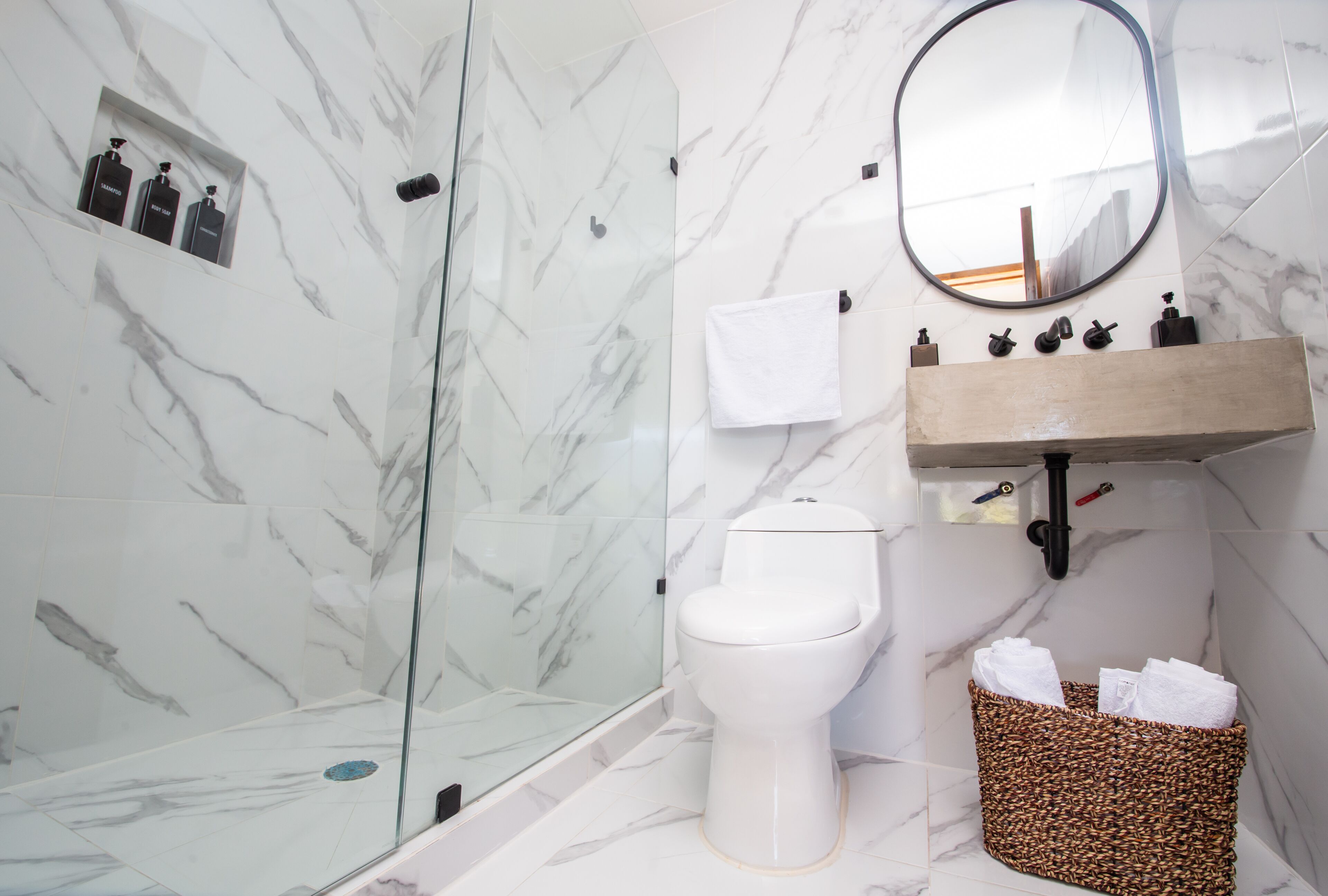 A bathroom with a marble wall, a glass shower enclosure, a toilet, a sink with soap dispensers, and a towel rack.