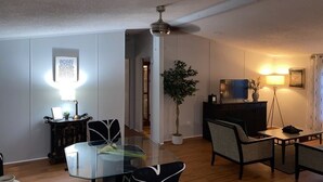Un salon moderne avec une table à manger en verre, deux chaises, un ventilateur de plafond, une plante en pot, un téléviseur et une lampe.