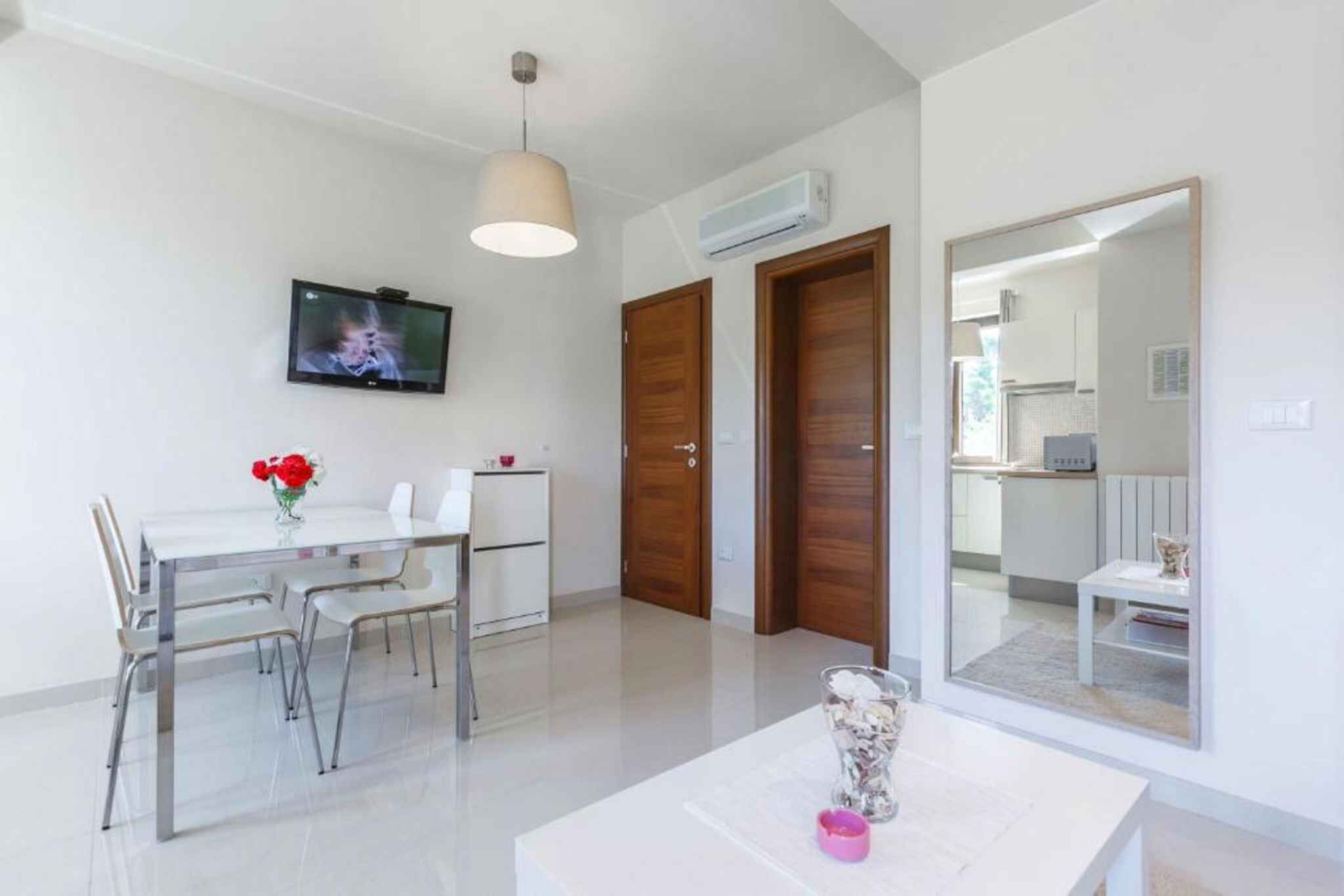 A modern, well-lit room with a dining table, chairs, a TV, and a large mirror reflecting the kitchen area.