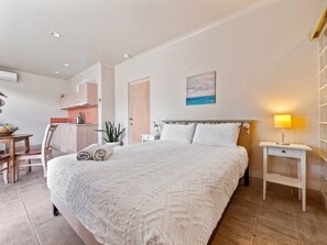 Un dormitorio con una cama grande, una mesita de noche con lámpara, un escritorio pequeño con silla y una kitchenette al fondo.