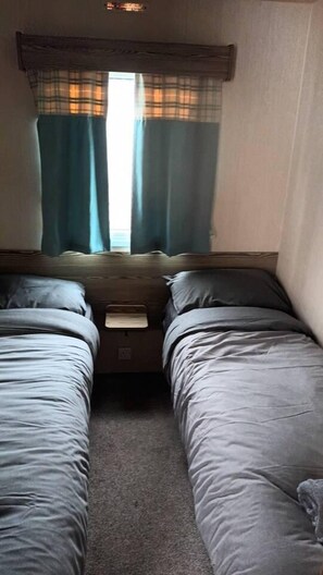 A small, dimly lit room with two single beds, a small table, and a window with curtains. - 3 bed Static 12ft Caravan Home From Home Somerset (Watchet)