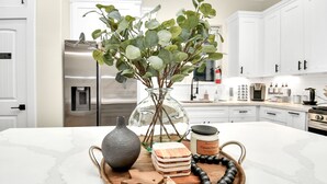 Eine Küche mit weißen Schränken, einem Edelstahl-Kühlschrank, einer Kaffeemaschine und einem Marmorarbeitsplatz mit einer Vase und grünen Blättern.