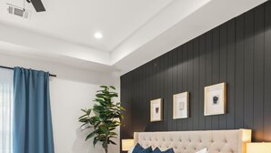 Ein modernes Schlafzimmer mit einer gesteppten Kopfteil, einem Bett mit weißen Leinen und vielen Kissen, einem Nachttisch mit Lampe, einem Deckenventilator und einem großen Fenster mit blauen Vorhängen.