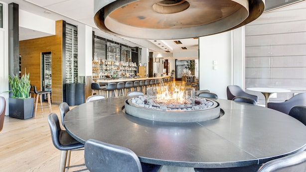 A modern bar area with a circular table featuring a central fire feature, surrounded by bar stools and a seating area with chairs.