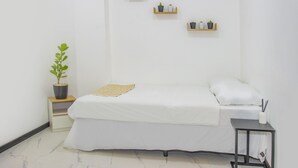 Une chambre à coucher minimaliste comprenant un lit, une petite table de chevet, une commode, une plante en pot et des étagères murales ornées d’objets décoratifs.