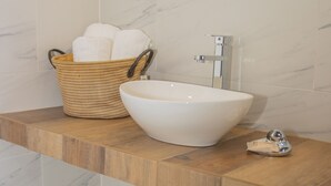 Une salle de bain avec un mur recouvert de carreaux de marbre, un plan de toilette en bois, un lavabo sur colonne blanc et un panier en osier contenant des serviettes.
