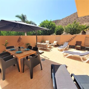 An outdoor patio with a dining table set for four, surrounded by lounge chairs and a large umbrella. - Très bel Appartement Lumineux 2chambres et Grande Terrasse à Palmmar (Palm Mar)