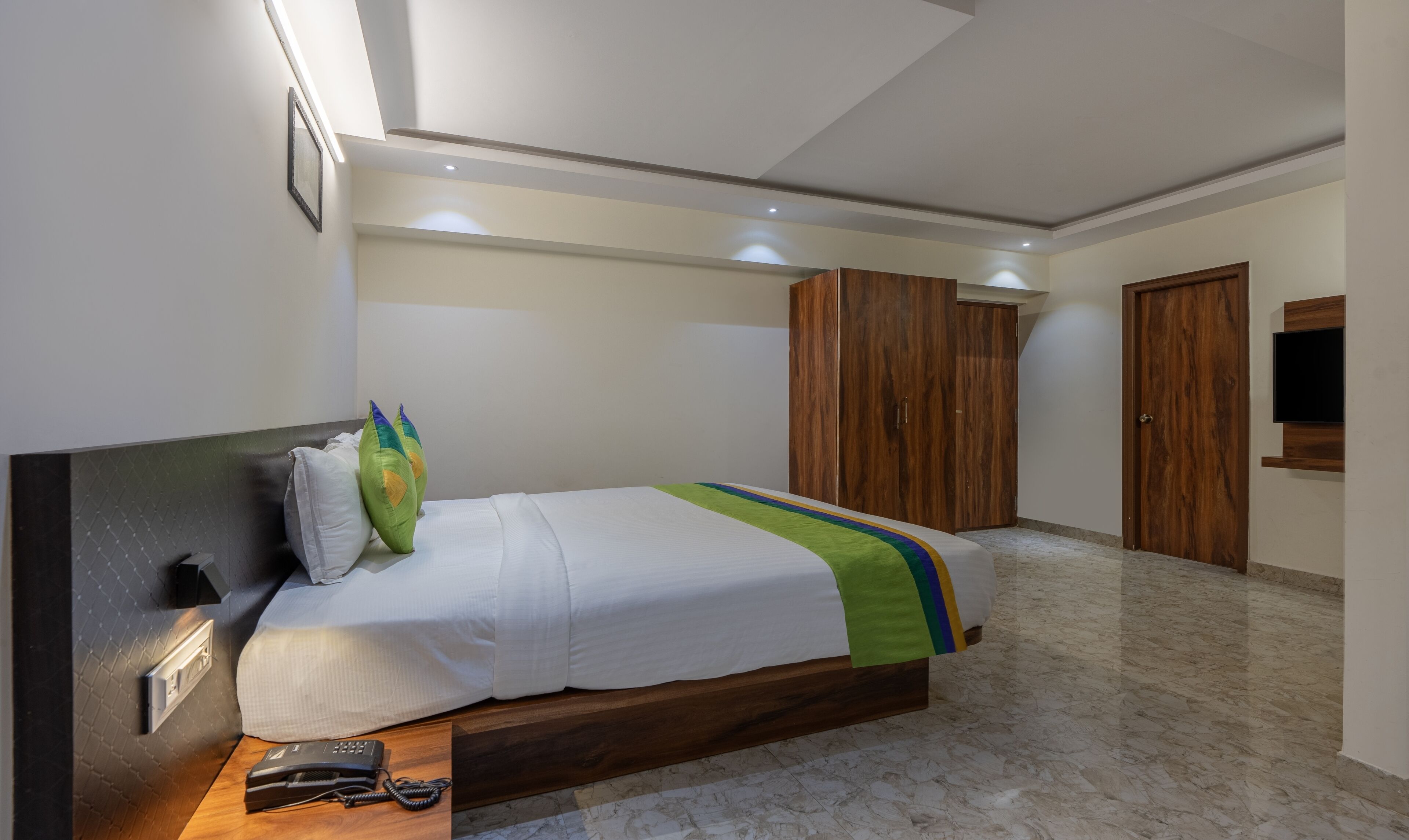A modern bedroom with a bed, a wooden wardrobe, a TV mounted on the wall, and a telephone on the bedside table.