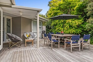 A covered patio with a wooden table and chairs, a grill, and a chair with a cushion. - Kohekohe Garden Cottage - Waikanae Holiday Home (Waikanae)