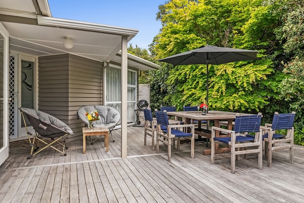 Une terrasse couverte avec une table et des chaises en bois, un barbecue et un fauteuil avec un coussin.
