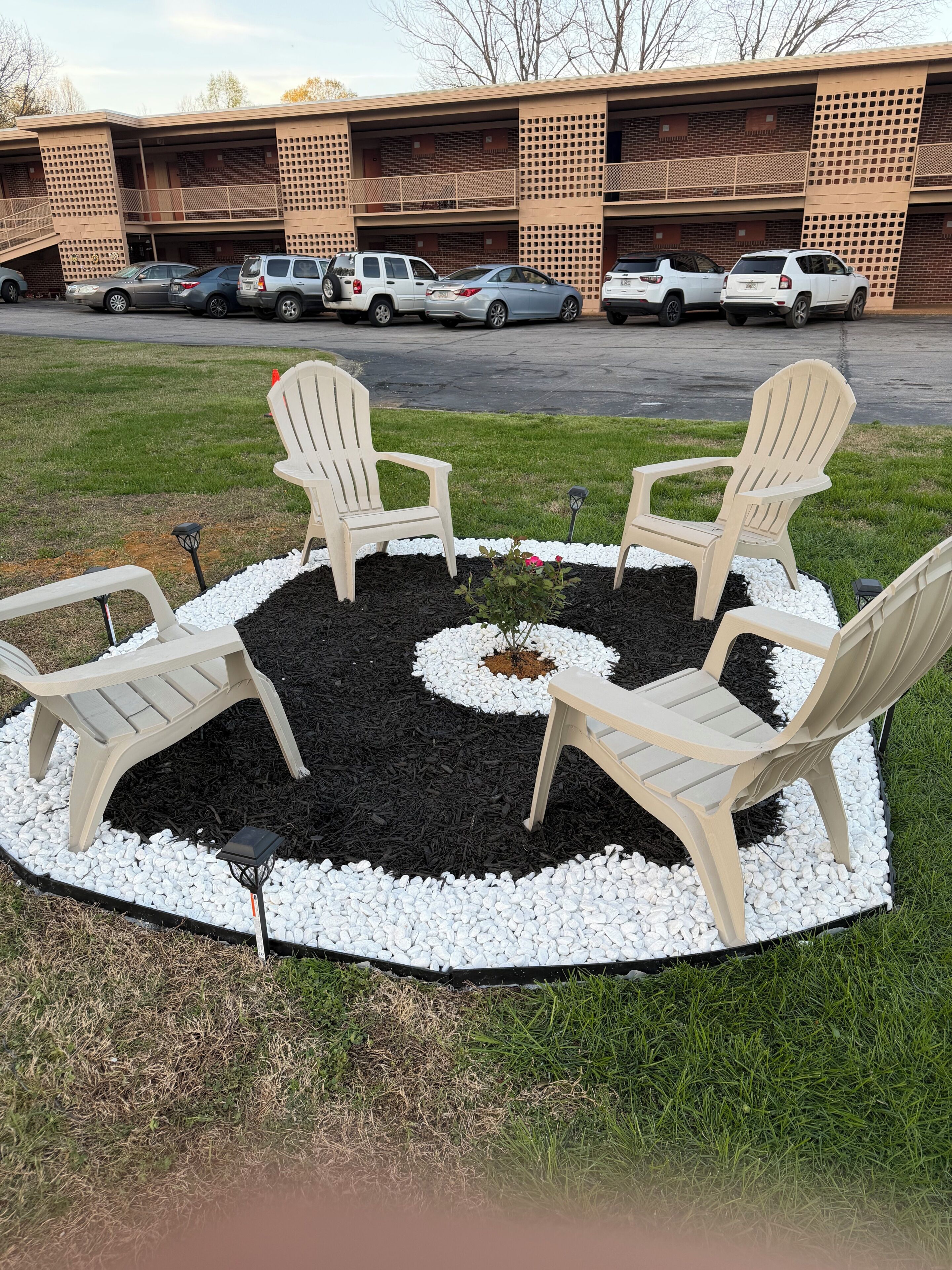 A circular seating area with four Adirondack chairs, surrounded by a gravel and mulch border, in front of a parking garage.