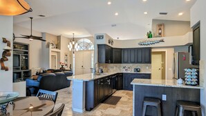 A modern kitchen with dark cabinets, a central island, and a dining area with a glass table and chairs. - Luxury south facing waterfront home with heated salt pool and spa! (Cape Coral)