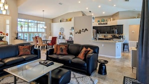 A spacious living room with a black leather sofa, a glass coffee table, and a ceiling fan. A kitchen with dark cabinets and a central island is visible in the background. - Luxury south facing waterfront home with heated salt pool and spa! (Cape Coral)