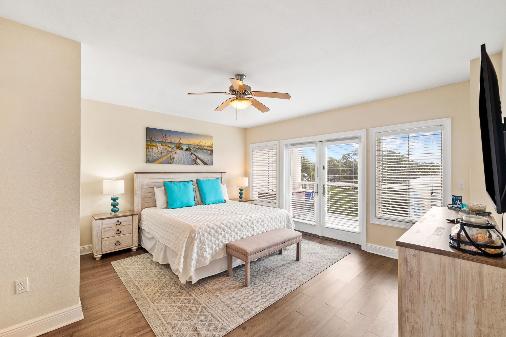 A bedroom with a bed, bedside tables, a bench, a ceiling fan, and large windows.