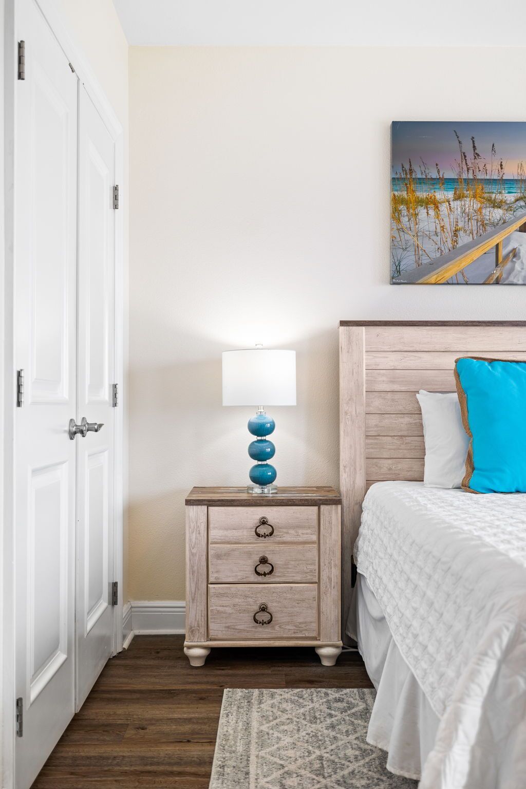 A bedroom with a wooden nightstand, a white lamp, a bed with white bedding, a blue pillow, and a painting of a beach scene on the wall.