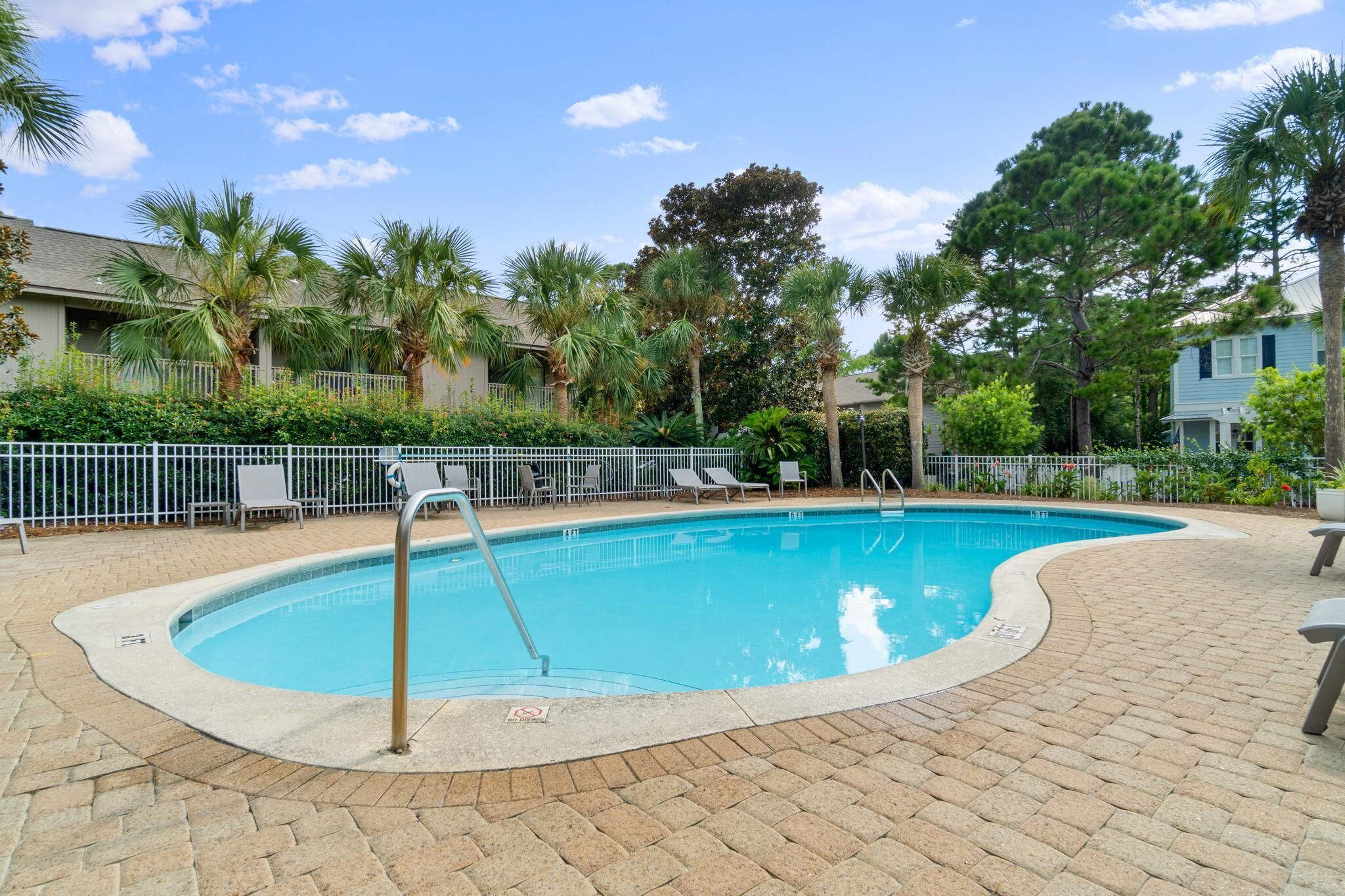 A swimming pool with a curved edge, surrounded by a brick patio, palm trees, and a fence with lounge chairs.