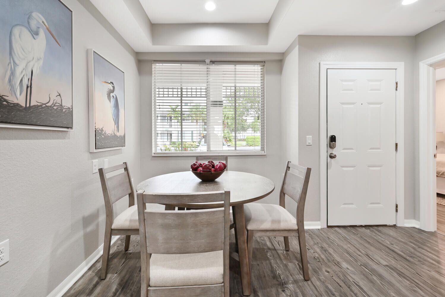 A dining area with a round table, four chairs, and a bowl of fruit. There are two windows with blinds, a white door, and artwork on the walls.