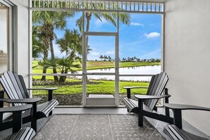 A balcony with two Adirondack chairs and a small table, overlooking a water body and palm trees. - Naples lake view condo at Lely golf resort! (Naples)