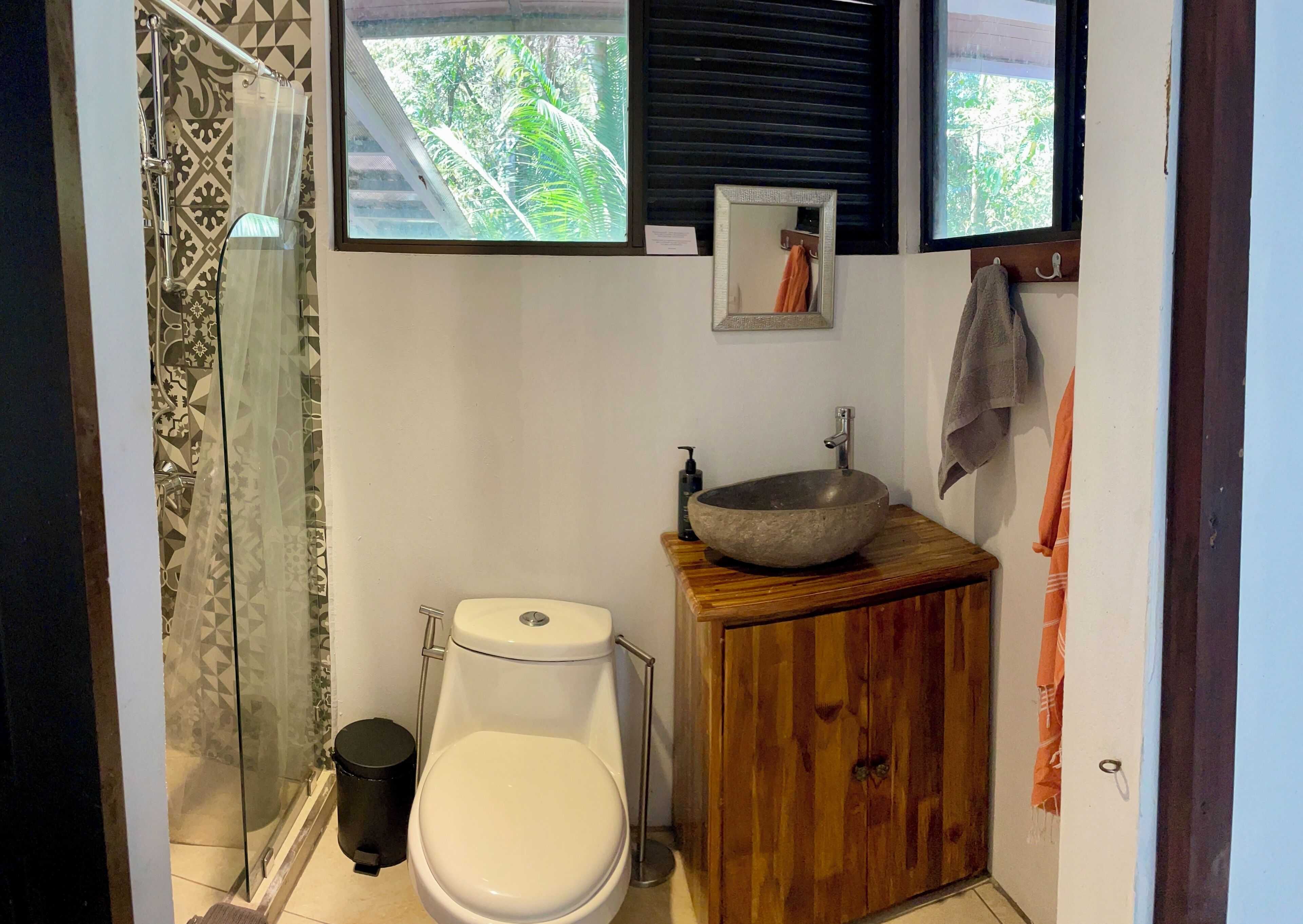 A bathroom with a toilet, a wooden cabinet with a sink, a black trash bin, and a shower with a patterned curtain.