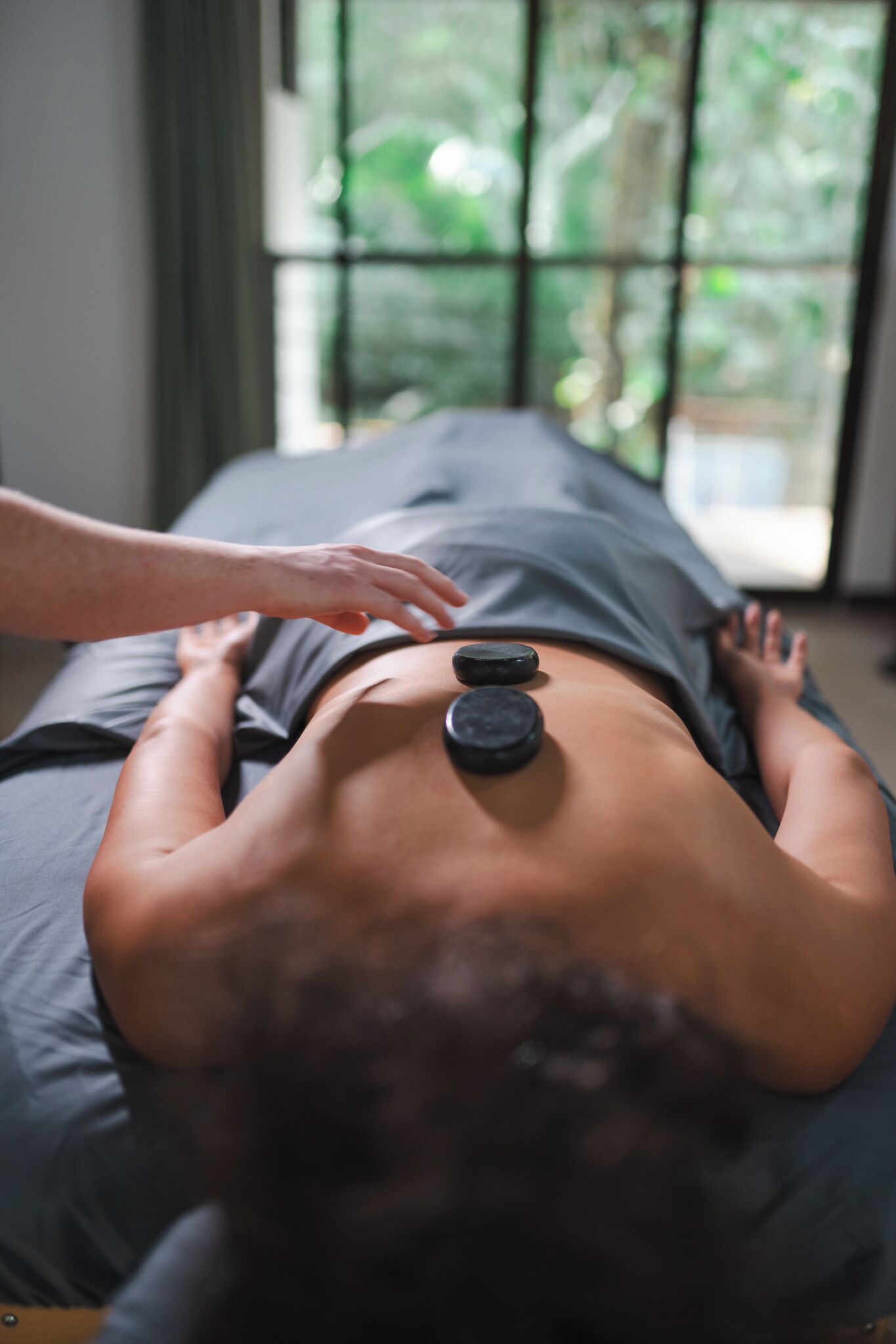 A person receiving a back massage with hot stones on a massage table in a room with a large window.
