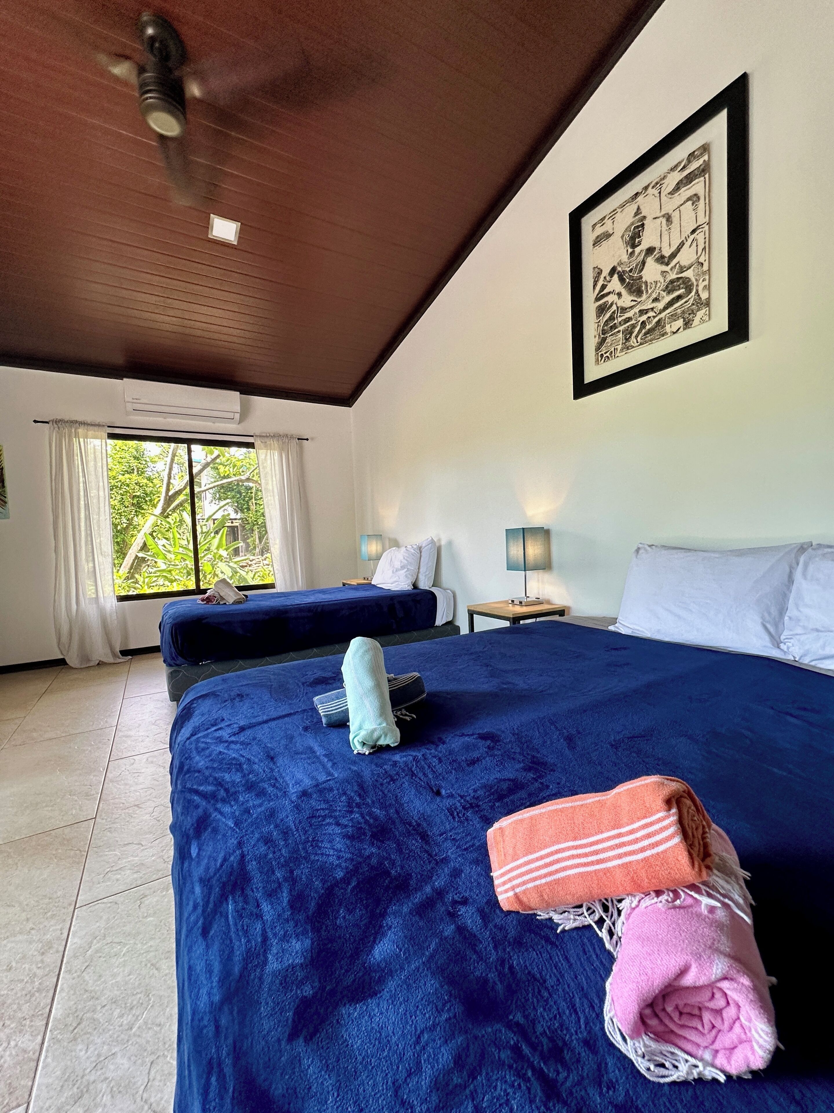 A hotel room with two beds, a ceiling fan, a large window with curtains, a framed artwork on the wall, and a small table with a lamp.