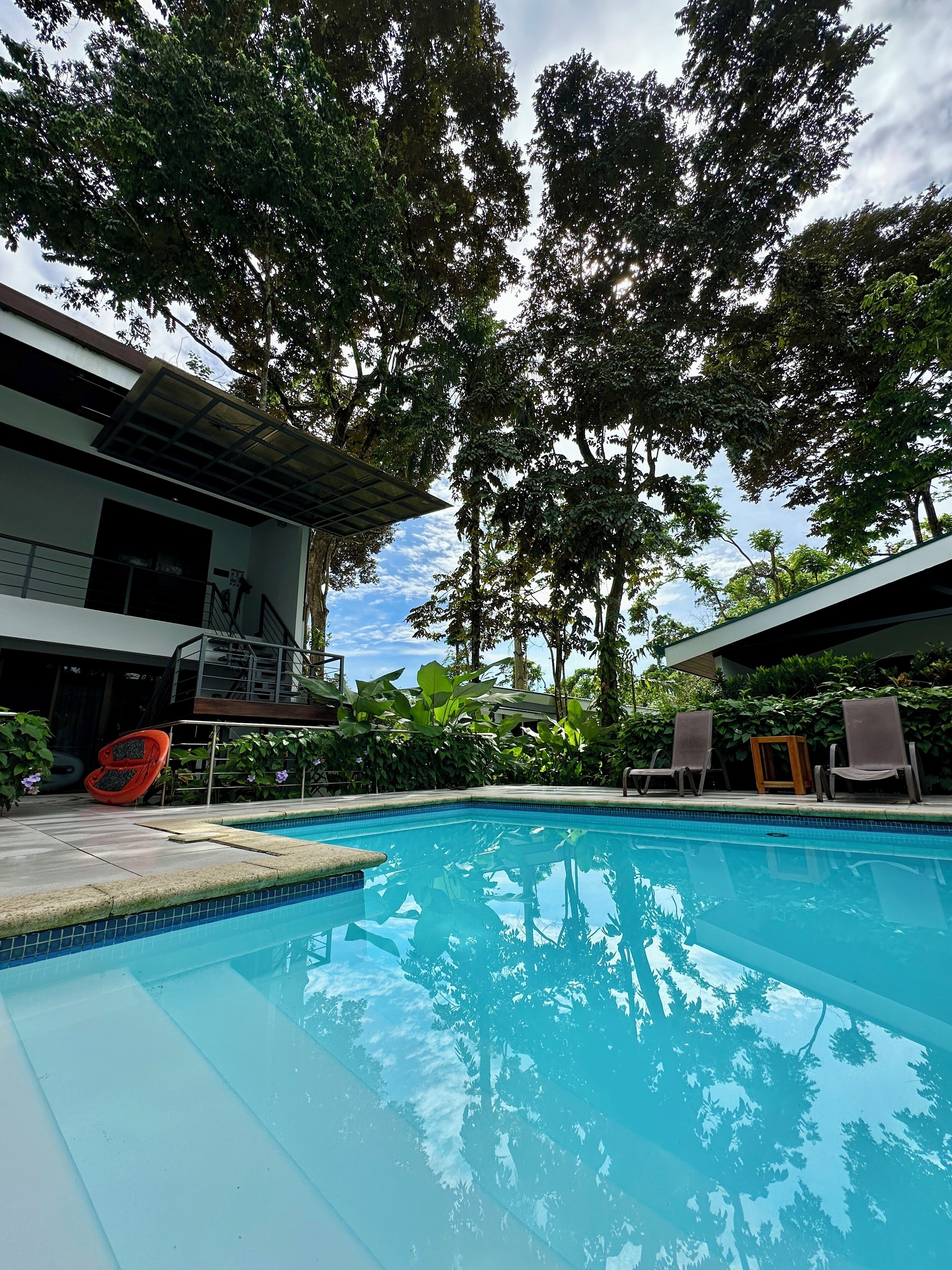 A swimming pool with a clear blue water surface, surrounded by lush green trees and a modern building with a balcony.