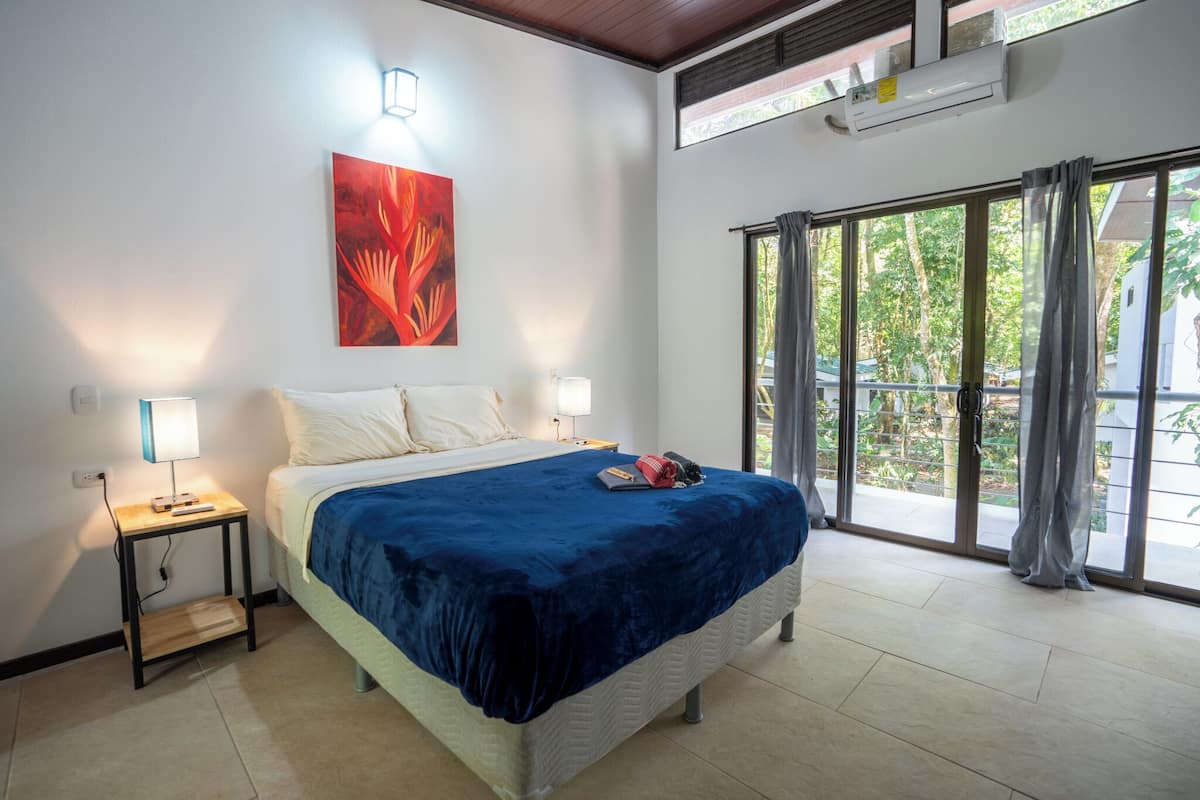 Ein Schlafzimmer mit einem großen Bett, einem Nachttisch, einer Wandlampe, einem Deckenventilator und einem großen Fenster mit Vorhängen.