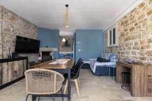 A room with a stone wall, a blue wall, a wooden dining table, a wicker chair, a sofa, and a television. - Villa Maraus- Seafront villa in Marausa, Sicily (Marausa)