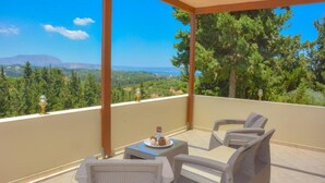 A balcony with wicker furniture, a small table with a plate of food, and a view of a mountainous landscape. - Villa Giorgos - Villa with private pool (Ágios Pávlos)