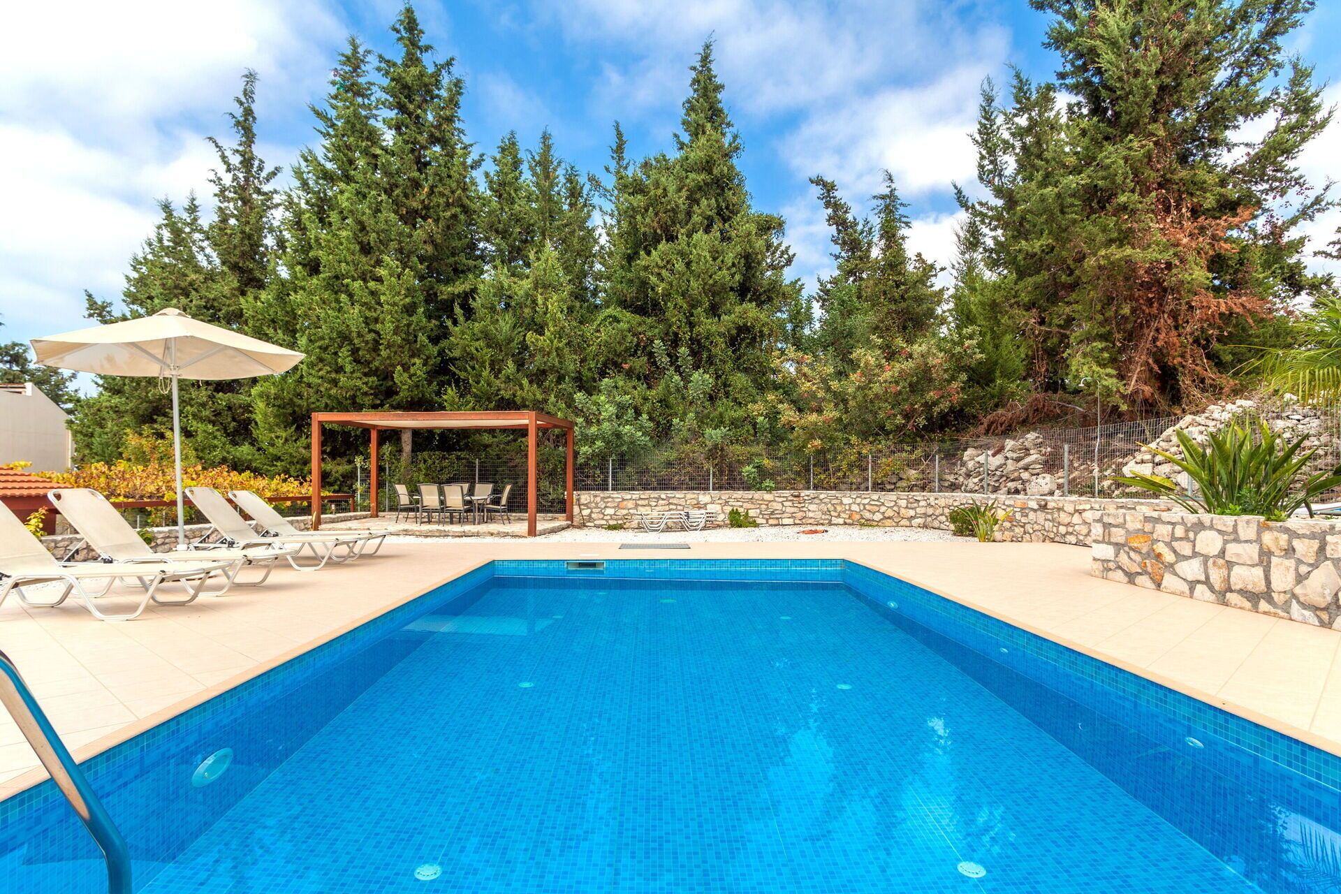 Una piscina con lettini e un pergolato, circondata da alberi e un muro in pietra.