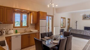 A modern kitchen with wooden cabinets, a dining area with a table set for two, and a view of the outdoors through large windows. - Villa Giorgos - Villa with private pool (Ágios Pávlos)
