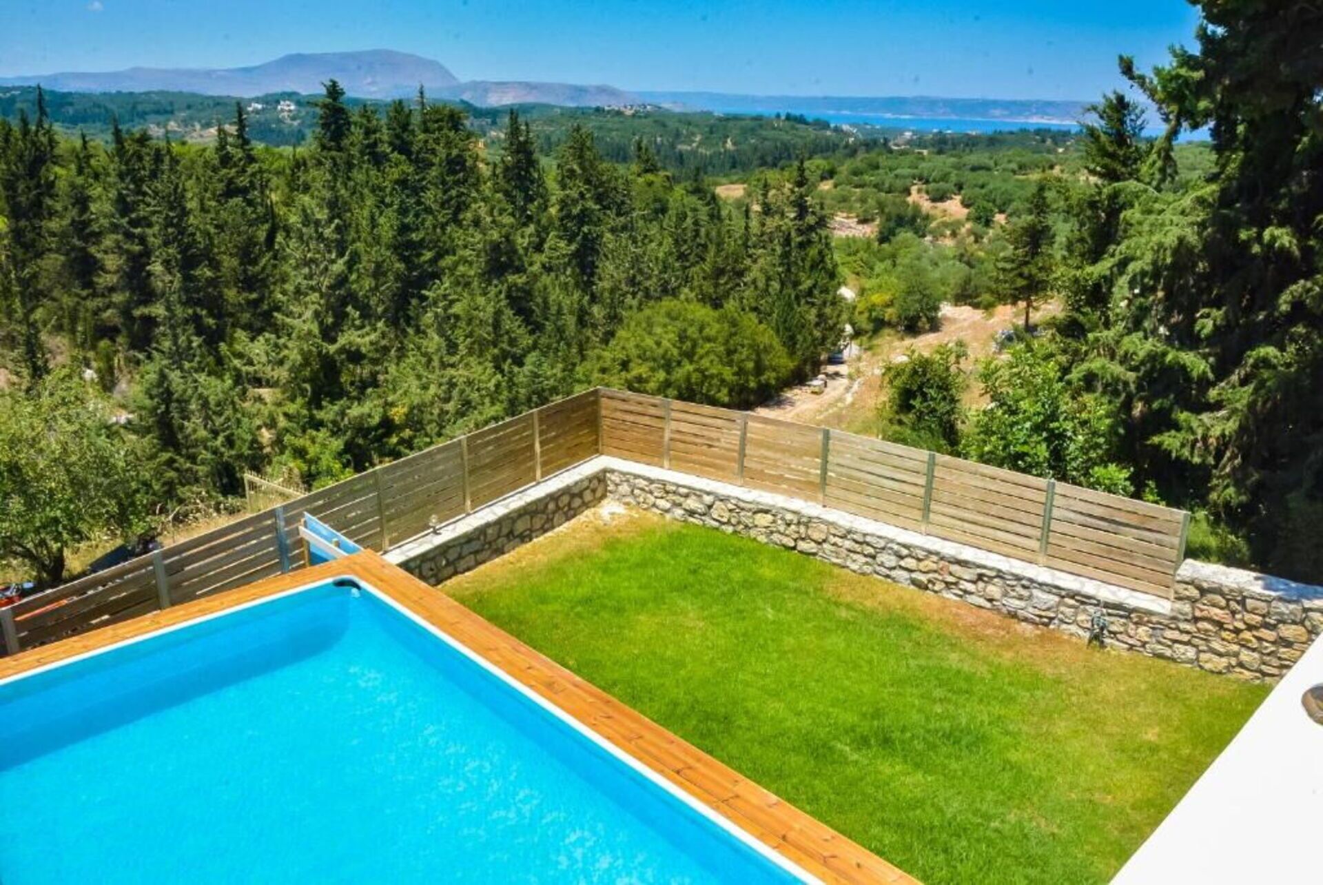 A swimming pool with a wooden deck and stone wall, surrounded by a grassy area and dense trees.